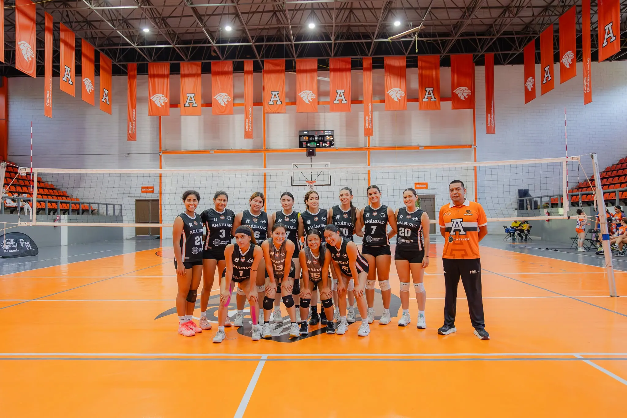 El equipo femenino de voleibol de la Universidad Anáhuac posa en su gimnasio, con los banderines del campus al fondo.