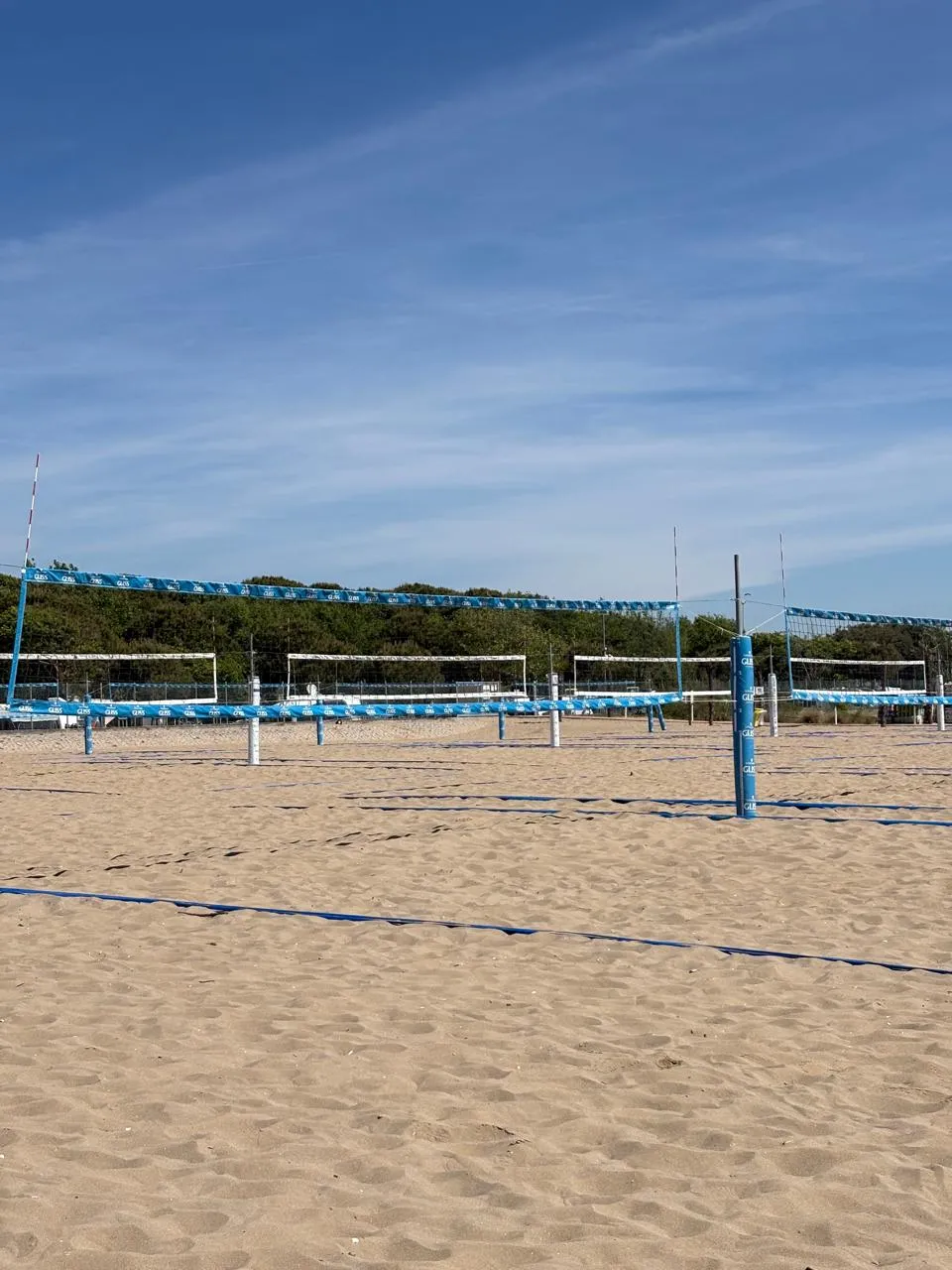 Canchas de voleibol de playa vacías bajo un cielo despejado, con los pinos del predio al fondo.